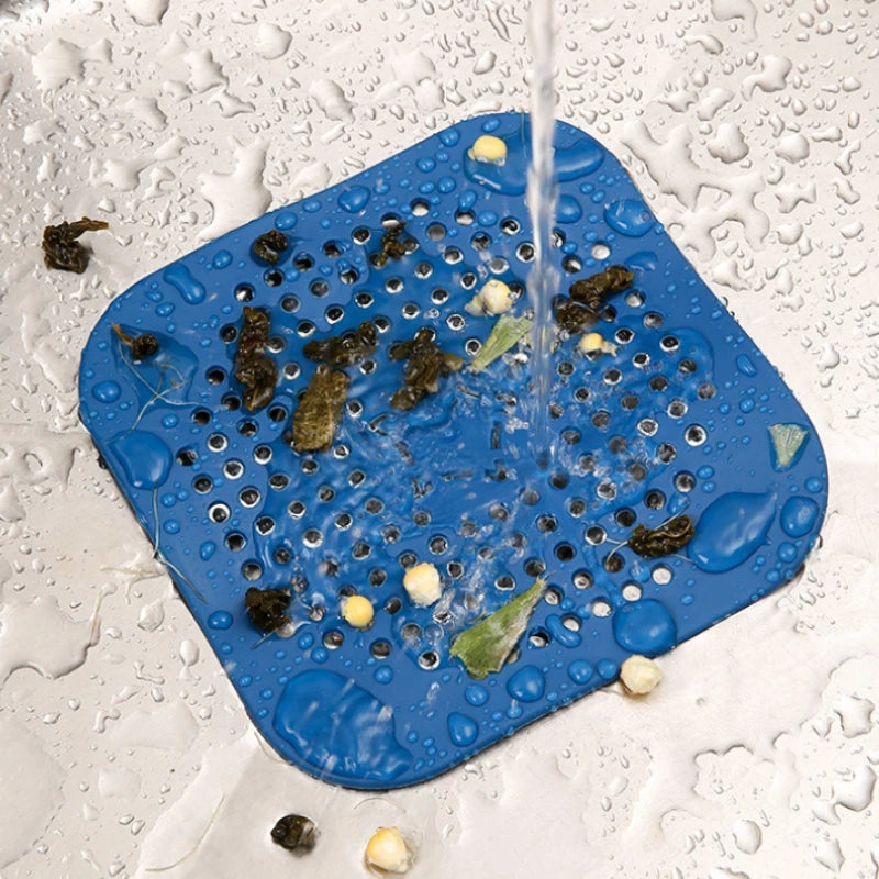 A blue square silicone drain filter mat with rounded corners and a grid of drainage holes, shown in a sink under running water with debris and soap suds falling through the mesh.