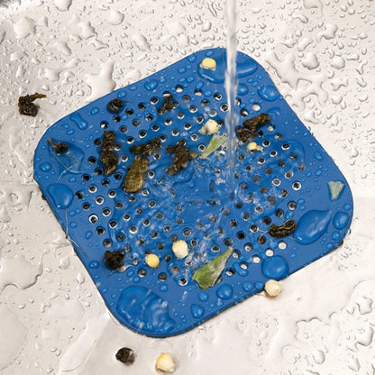 A blue square silicone drain filter mat with rounded corners and a grid of drainage holes, shown in a sink under running water with debris and soap suds falling through the mesh.