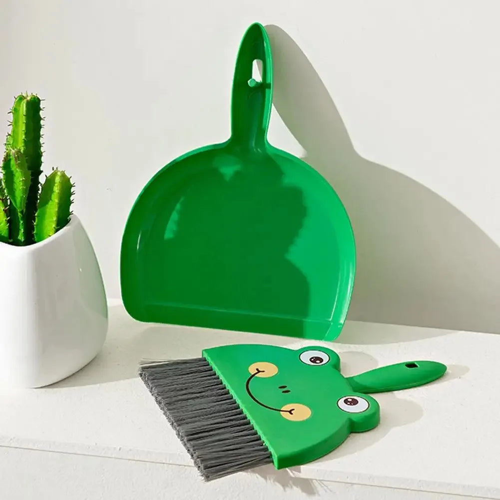 Green dustpan and brush set with a face design on a white surface next to a potted cactus.