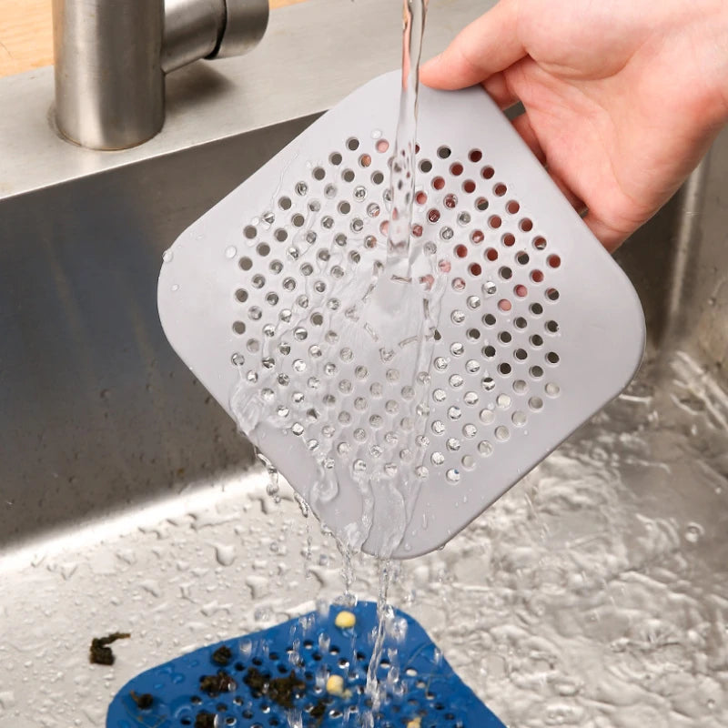 Gray square sink strainer being rinsed under running water in a kitchen sink.