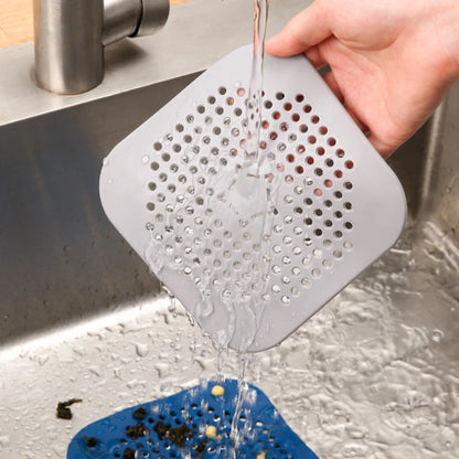 Gray square sink strainer being rinsed under running water in a kitchen sink.