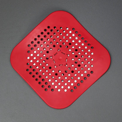 A red silicone or rubber drain grommet with a diamond-shaped body, rounded corners, and a circular pattern of pre-drilled holes on a dark gray background.