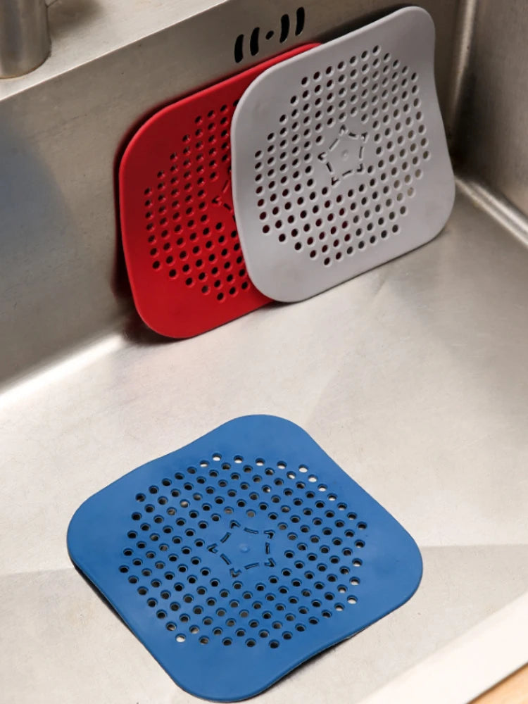 Three square drain stoppers with rounded corners in red, gray, and blue, shown inside a stainless steel kitchen sink. Each stopper has a dense grid of circular holes and a small star-like cutout in the center.