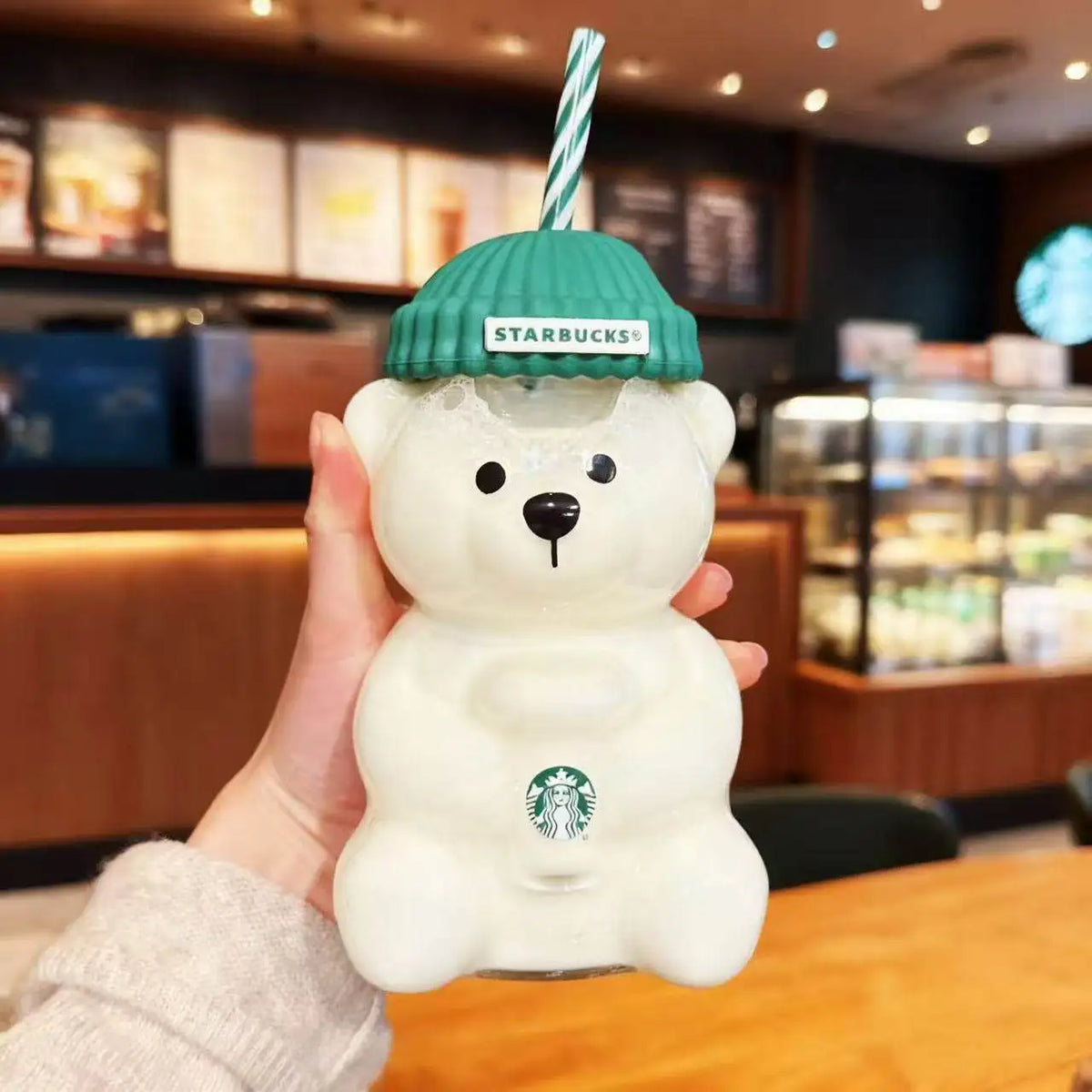 Starbucks bear-shaped cup with green knit hat held in a hand inside a Starbucks store.