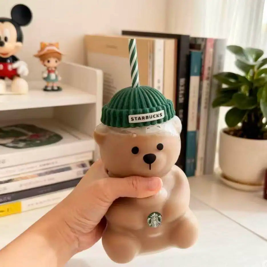Starbucks bear-shaped cup with a green lid held by a hand, with a bookshelf and plant in the background.